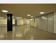 Unterirdischer Fußgängertunnel mit gefliesten Wänden und Boden, mehreren Wandleuchten und einer schwarzen Säule links im Vordergrund. An den Wänden hängen Werbeplakate und ein Übersichtsplan.