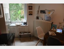 Kleine Küche mit einem Fenster, das auf eine von Efeu bewachsene Wand blickt. Auf der Fensterbank stehen Glasgefäße und eine Kaffeemaschine, während unter dem Fenster ein Futternapfhalter steht; ein Heizkörper, ein kleiner Esstisch mit Stuhl und Bücher sowie Dekorationen sind sichtbar.