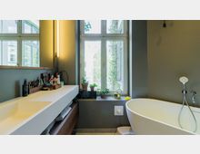 Modernes Badezimmer mit einem großen Fenster und einer freistehenden Badewanne. Links befindet sich ein breiter Waschbeckenbereich mit Ablage, Spiegel und beleuchtetem Wandstreifen, während die Fensterbank mit Pflanzen und kleinen Gegenständen dekoriert ist.