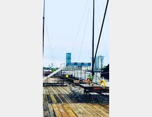 Holzdeck eines Schiffes mit Tischen und Stühlen im Vordergrund, umgeben von Takelage und Seilen. Im Hintergrund sind moderne Gebäude mit Glasfassaden und der Fluss sichtbar.