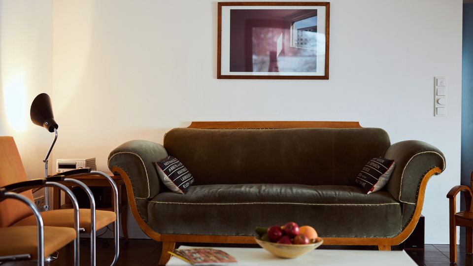 A cozy living room with a green upholstered vintage sofa flanked by wooden armrests, a small side table with a modern lamp, and orange chairs with chrome frames. A framed photograph is mounted on a white wall above the sofa, and a bowl of fruit sits on a white coffee table in the foreground.