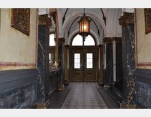 Eingangsbereich eines historischen Gebäudes mit verzierten Wänden in schwarz-goldenem Marmordekor, einem gemusterten Fliesenboden und einer großen Holztür mit verglasten Fenstern im Hintergrund. Über der Tür hängt eine an Ketten befestigte Laterne, und an den Seiten befinden sich dekorative Spiegel mit goldenen Rahmen.