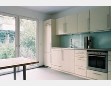 Moderne Küche mit weißen Schränken, einer durchgehenden Arbeitsplatte und einer Glasrückwand in hellblau. Links befinden sich bodentiefe Fenster mit Blick auf einen Garten, rechts eine eingebaute Spüle, Backofen und Kaffeemaschine.