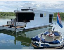 Ein modernes Hausboot mit weißem Rumpf und großen Fenstern liegt an einem Anlegesteg auf einem ruhigen See, umgeben von bewaldeter Uferlandschaft. Im Vordergrund ist ein kleines Motorboot mit einer niederländischen Flagge zu sehen.
