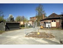 Ehemaliges Industriegelände mit roten Backsteingebäuden, teilweise mit zerbrochenen Fenstern und geschlossenen Türen. Im Vordergrund ein Zaun mit Baustellenabsperrung, ein kleiner Schuppen und eine gepflasterte Straße, umgeben von Bäumen und Sträuchern.