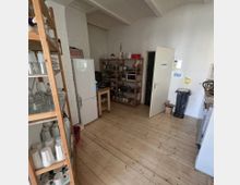 Kleine Küche mit Holzboden und weiß gestrichenen Wänden. Zu sehen sind offene Holzregale mit Geschirr und Küchenutensilien, ein Kühlschrank, eine Mülltonne sowie eine offene Tür mit der Aufschrift „Backstage“.