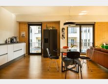 Moderne Wohnküche mit Holzboden, einer weißen Einbauküche links, und einem Essbereich mit einem runden Tisch und unterschiedlichen Stühlen in der Mitte. Große Fenster mit Balkonzugang und hellem Holz an den Wänden lassen viel Tageslicht einfallen, während im Hintergrund eine braune Ledercouch steht.