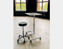 Ein leerer Raum mit Betonboden und weißen Holzrahmenfenstern, durch die grüne Bäume sichtbar sind. Im Vordergrund stehen ein höhenverstellbarer Hocker auf Rollen und ein kleiner, mobiler Stehtisch mit Metallgestell.