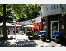 Eine kleine Ladenzeile mit Backsteingebäuden und Geschäften, darunter ein Imbiss und Souvenirshops, umgeben von außen aufgestellten Tischen, Sonnenschirmen und Sitzgelegenheiten unter Bäumen. Im Vordergrund sind dekorative Holzbänke und Fässer platziert.