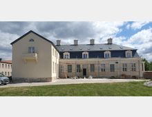 Ein mehrstöckiges Gebäude mit heller, beige-gelber Fassade und dunklem Schieferdach, das sich offensichtlich in Renovierung befindet. Vor dem Gebäude sind Gerüste angebracht, und die Umgebung zeigt eine gepflasterte Fläche und eine Rasenfläche unter einem bewölkten Himmel.