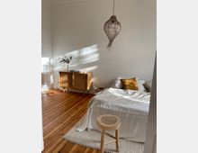 Helles Schlafzimmer mit Holzboden, einem Bett mit weißer Bettwäsche, einem goldfarbenen Kissen, einem runden Holzhocker und einem gewebten Teppich. Im Hintergrund stehen ein Sideboard aus Holz mit Korbelementen und eine Vase mit Zweigen sowie eine geflochtene Hängeleuchte an der Decke.