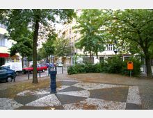 Kleine städtische Platzszene mit gepflastertem Boden in Mosaikmustern, umgeben von Bäumen und Sträuchern. Im Hintergrund befinden sich Wohnhäuser mit Balkonen, geparkte Autos und eine orangefarbene Abfalltonne, mit einer dekorativen, gusseisernen Wasserpumpe im Vordergrund.