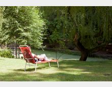 Eine Person liegt auf einem roten Liegestuhl auf einer Wiese neben einem Teich, der von grünen Bäumen und einem großen Baum mit tief hängenden Ästen umgeben ist. Im Hintergrund sind schattige Waldstücke und Sitzbänke zu erkennen.