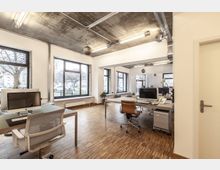 Modernes Büro mit großen Fenstern, Holzfußboden und sichtbarem Beton an der Decke, ausgestattet mit Schreibtischen, Bürostühlen und Computerarbeitsplätzen. Im Hintergrund sind mehrere Bäume und parkende Autos durch die Fenster sichtbar.