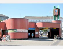 Eingang zur U-Bahn-Station Fehrbelliner Platz in Berlin mit geschwungenen roten Fassaden, gelben Deckenelementen und einem grünen Säulenaufbau mit U-Bahn-Schild. Im Vordergrund steht ein Imbiss mit Preistafel, und Fahrräder sind an einem Geländer abgestellt.