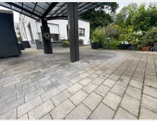 Gepflasterter Hof mit einem modern überdachten Carport und einem weißen Gebäude im Hintergrund. Rechts befinden sich Pflanzkübel mit verschiedenen Blumen und Sträuchern, die an einen Garten grenzen.