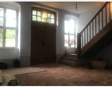 Ein rustikaler Eingangsbereich mit grau verputzten Wänden, einem großen Holztor in der Mitte, zwei großen Fenstern mit Sprossen auf beiden Seiten und einer Holztreppe, die nach oben führt. Der Boden besteht aus roten Terrakottafliesen, und die Fenster lassen Tageslicht herein.