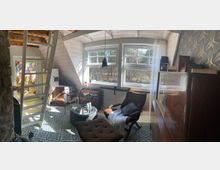 Gemütlicher Dachwohnraum mit großen, weißen Sprossenfenstern, durch die Tageslicht einfällt und den Blick auf einen Wald freigibt. Im Raum befinden sich eine Holzleiter, eine dekorative Tapetenwand im goldenen Muster, ein Sessel mit Hocker, ein Glastisch sowie ein Bücherregal und weitere Holzmöbel.