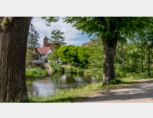 Ein Fluss verläuft durch eine grüne Parklandschaft mit Bäumen und Rasenflächen. Im Hintergrund sind rote Ziegeldächer, ein Kirchturm und weitere Gebäude sichtbar.