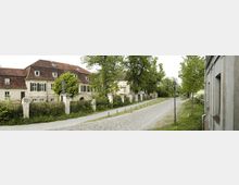 Eine gepflasterte Straße mit Laternen führt vorbei an historischen Gebäuden mit roten Ziegeldächern und verputzten Fassaden. Im Vordergrund sind eine alte Steinmauer mit Pfosten und zahlreiche Bäume sowie Grünflächen zu sehen.