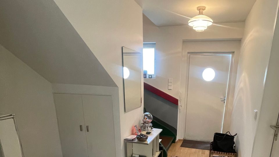 Schmaler Eingangsflur mit hellem Holzboden, weißer Decke und Deckenleuchte, der zu einer weißen Haustür mit rundem Bullaugenfenster führt. Links stehen ein Einbauschrank unter der Treppe und ein kleiner Konsolentisch mit Spiegel, rechts eine Sitzbank und ein langer Läuferteppich entlang des Gangs.