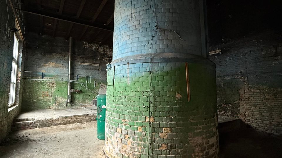 Innenraum eines verlassenen Industriegebäudes mit großem zylindrischem gemauertem Schornstein oder Ofen in der Mitte, unten grün gestrichen und oben grau. Umgeben von rauen Backsteinwänden, Betonboden mit Schmutz und Rissen sowie hohen Fenstern an der linken Seite unter einem Holzdach.