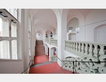Historischer Treppenaufgang mit weißen Wänden, gewölbter Decke und großen Fenstern, die Tageslicht hereinlassen. Die Treppe aus rotem Bodenbelag wird von weißen Balustraden mit kunstvollen Verzierungen und Laternenleuchten flankiert.
