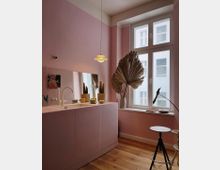 Kleine Küche mit rosa Wänden und minimalistischer Einrichtung, darunter eine flache rosa Küchenzeile mit integriertem Waschbecken und verschiedenen Accessoires wie Pflanzen in Töpfen, Geschirr und Dekorationen. Ein großes Fenster mit weißem Rahmen lässt Tageslicht herein, daneben steht eine Palme in einer Vase; der Boden besteht aus heller Holzvertäfelung.