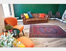 Wohnzimmer mit Holzboden, einer ledernen Chesterfield-Couch, bunten Kissen, einem dunkelgrünen Wandhintergrund und einem großen, roten Orientteppich. Im Vordergrund stehen orangefarbene Stühle und ein kleiner Tisch mit einer Topfpflanze.