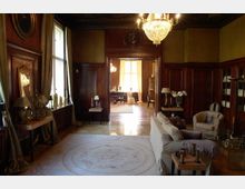 Ein elegant eingerichteter historischer Salon mit Holzwänden, großen Fenstern und einem Kronleuchter an der Decke. Im Vordergrund befinden sich ein Sofa, ein Teppich und dekorative Möbel, im Hintergrund ein angrenzender Raum mit weiteren Sitzgelegenheiten und großen, lichtdurchfluteten Fenstern.