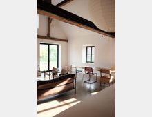 Ein heller Innenraum mit hohen Decken, Sichtbalken und großen Fenstern. Der Raum ist sparsam eingerichtet mit Stühlen, einem Sofa, einem Tisch und modernen Designmöbeln auf einem polierten Betonboden.