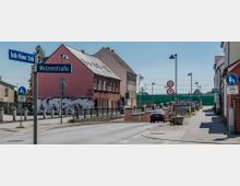 Straßenszene mit Abzweigung zwischen der Wolzenstraße und der Großen Milower Straße, flankiert von Gebäuden mit teils mit Graffiti bemalten Wänden. Im Hintergrund sind Bahngleise und eine grüne Schallschutzwand zu sehen, sowie Verkehrsschilder und Gehwege mit Laternen.