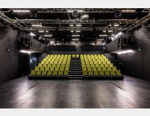 Innenraum eines schwarzen Theater- oder Studiorsaals mit ansteigender Tribüne aus hellgrünen Sitzreihen und mittigem Treppenaufgang. Über der Bühne hängen Metalltraversen mit Scheinwerfern und Technik, vor der Tribüne liegt eine freie, dunkle Spielfläche.