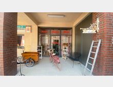 Eingangsbereich eines kleinen Geschäfts mit Glasfront, auf der die Wörter "Deko", "Blumen" und "Möbel" stehen. Vor dem Geschäft stehen ein rotes Fahrrad, ein Lastenrad mit der Aufschrift "Kiez Bike", und ein paar Regale, daneben eine Leiter und ein dekoratives Schild mit der Aufschrift "Blumen".