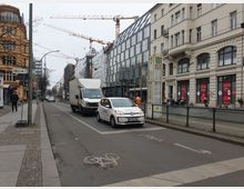 Straßenszene mit Wohn- und Geschäftshäusern, darunter ein modernes verglastes Gebäude und ein älteres Gebäude mit heller Fassade. Im Vordergrund sind eine Fahrbahn mit Autos und ein Fahrradweg, dazu eine Bushaltestelle mit gelbem Schild sichtbar; im Hintergrund erheben sich Baukräne.