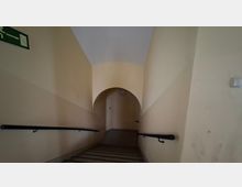 Schmales Treppenhaus mit hellbeigen Wänden und Handläufen an beiden Seiten, das nach oben zu einem rundbogigen Durchgang führt. Die Decke wölbt sich leicht, und am Ende des Aufgangs ist eine Tür im Halbrund sichtbar.