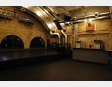 Innenaufnahme einer Veranstaltungsstätte mit Backsteinmauern, schwarzer Bühne und großen, bogenförmigen Fenstern. Sichtbare Metall-Lüftungsschächte sowie Licht- und Tontechnik hängen an der Decke; rechts im Bild ist eine Bar mit Getränkespendern zu sehen.