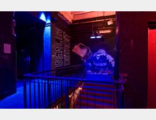 Industrieller Treppenhausbereich mit schwarzer Wand, auf der in weißer Schrift "Berlin Detroit" steht, sowie ein Wandgemälde eines Gorillas und eines Ghettoblasters. Die Szene ist in blaues und rotes Licht getaucht, mit sichtbaren Metallgeländern und Kabeln an der Decke.