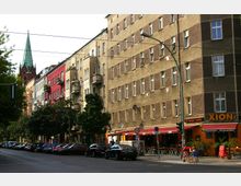 Straßenszene in einer Stadt mit mehrstöckigen Wohngebäuden, die teils farbig gestrichen sind, Balkonen und großen Fenstern. Im Vordergrund steht eine Straße mit geparkten Autos, während ein Café mit roten Markisen und Menschen, die draußen sitzen, sichtbar ist; im Hintergrund ragt ein Kirchturm mit grünem Dach empor.