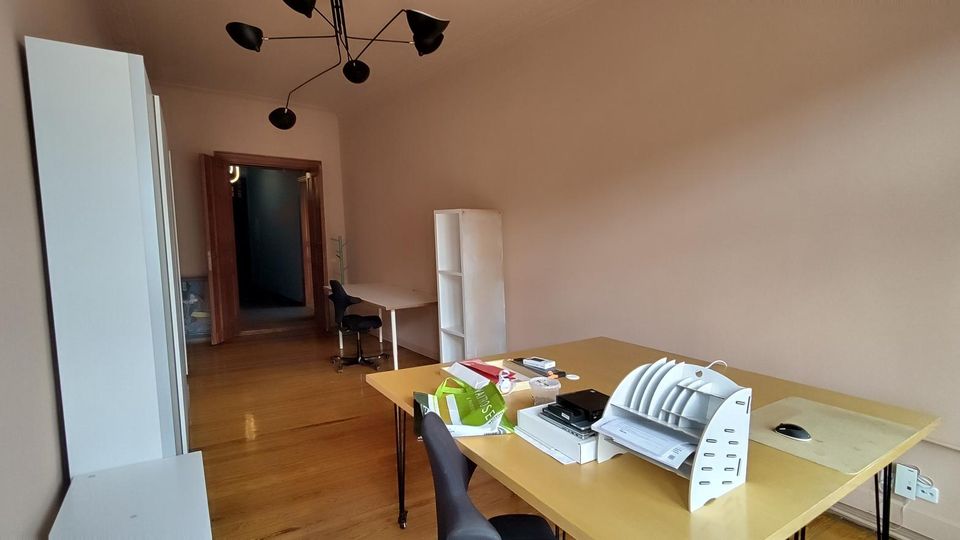 Schmales Büro mit Holzfußboden, hellen Wänden und moderner Deckenleuchte; links stehen hohe weiße Schränke, rechts ein großer Schreibtisch mit Ablagen. Im Hintergrund sind ein weiterer Arbeitsplatz, ein Regal und eine offene Tür zu einem Flur zu sehen.