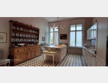 Helle, großzügige Wohnküche mit schwarz-weißem Fliesenboden, zentraler Kücheninsel und hohen Fenstern. Links steht ein großer Holzbuffetschrank mit offenem Tellerregal, rechts eine weiße Einbauküche mit Arbeitsplatte und Oberschränken.