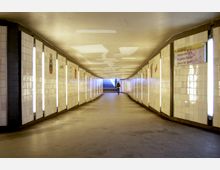 Unterirdischer Fußgängertunnel mit weißen Fliesenwänden, vertikalen Leuchtstreifen und farbigen Wandzeichnungen. Im Hintergrund führen Treppenstufen ins Freie, eine Person geht durch den Tunnel.