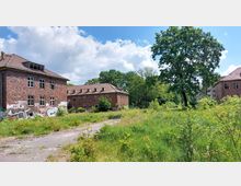 Alte Backsteingebäude mit Graffiti auf den Wänden, umgeben von einer verwilderten Wiese und vereinzelten Bäumen unter einem sonnigen Himmel. Im Vordergrund führt ein verwitterter Gehweg durch das Gelände.