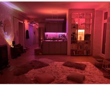 Wohnzimmer mit offener Küche in warmem rosa und orangefarbenem Stimmungslicht. Der Raum enthält ein flauschiges Teppicharrangement mit Kissen, eine Bücherwand, eine Küchenzeile mit eingebautem Licht und dekorative Elemente wie Spiegel und Pflanzen.