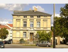 Historisches zweigeschossiges Wohnhaus mit gelber Fassade, dekorativen Stuckelementen und einem großen Holzeingang. Vor dem Gebäude stehen parkende Autos, und der Vorgarten ist mit einem Zaun und Bäumen eingefasst.
