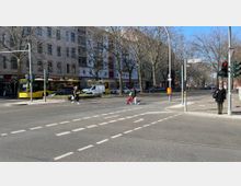 Straßenkreuzung in einer Stadt mit Fußgängerüberwegen, Ampeln und mehreren Passanten, darunter eine Person mit einem kleinen Hund. Im Hintergrund befinden sich mehrstöckige Wohnhäuser, Geschäfte im Erdgeschoss und ein gelber Linienbus an einer Haltestelle.