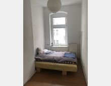 Schlichtes Schlafzimmer mit weiß gestrichenen Wänden, einem kleinen Fenster mit Blick auf ein gegenüberliegendes Gebäude und einem einfachen Bett mit Bettdecke und Kissen. Eine Papierlampe hängt von der Decke, und unter dem Fenster befindet sich eine Heizung.