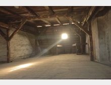 Dunkler Dachboden mit Holzbalkenkonstruktion und einer hölzernen Bodenfläche. Im Hintergrund fällt Licht durch ein kleines Fenster, das teilweise mit Dachziegeln umgeben ist.