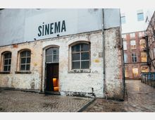 Backsteinfassade eines alten Gebäudes mit der Aufschrift „SINEMA“ über den Fenstern. Ein geöffneter Eingang mit einem roten Vorhang und Blick auf warmes Innenlicht; gepflasterter Hof mit weiteren Backsteingebäuden im Hintergrund.