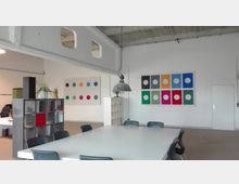 Ein moderner Besprechungsraum mit einem großen weißen Tisch, umgeben von schwarzen Stühlen. An den weißen Wänden hängen abstrakte Kunstwerke mit bunten Kreisen, darüber eine hängende, industrielle Lampe; im Hintergrund ein offenes Regal und eine geschlossene Tür.